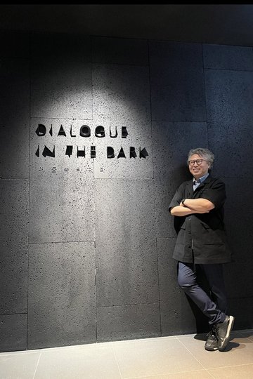 Photo of Yong-hee Song infront of a wall with the lettering "Dialogue in the Dark" in English and Korean.