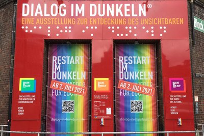 Foto der Eingangstüren des Dialoghauses Hamburg, die mit regenbogenfarbenen Banner geschmückt sind, auf welchen die Wiedereröffnung zum 02.07.2021 angekündigt wird.