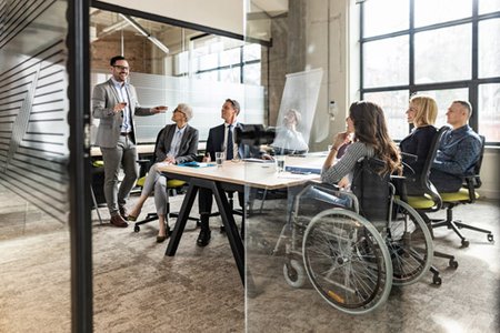 Foto einer gruppe von Mitarbeitern in einem Besprechungsraum. Ein Kolleg steht vor der Gruppe und erklärt etwas. Eine Frau sitzt im Rollstuhl.