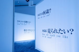 Foto des Tunnels der Fragen aus der früheren Dialog mit der Zeit-Ausstellung in Tokio: auf weißen Wänden entlang des Tunnels stehen Fragen in englischer und japanischer Schrift, wie z.B. "Wie alt bist du?" oder "Wie alt möchtest du geschätzt werden?".