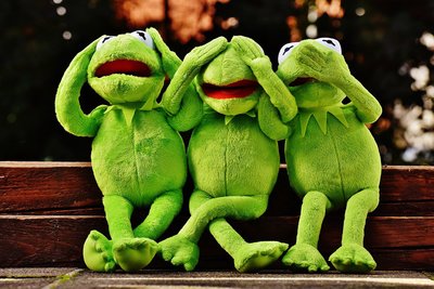 A photo of three green stuffed frog puppets, sitting next to each other, one covers his ears, the second his eyes and the third his mouth.n