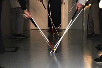 Foto von einigen Personen, von denen nur die Beine gezeigt werden, die mit Hilfe eines Langstocks einen Korridor entlang gehen. 