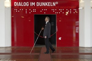 Picture of a guide with a white cane passing by the entrance of the Dialogue in the Dark exhibition in Hamburg