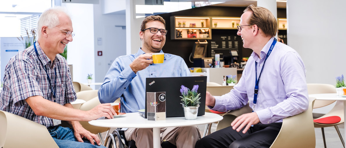 Foto von drei Männern, die zusammen an eienm Tisch sitzen, einer von ihnen im Rollstuhl. Während sie Kaffee trinken, schauen sie etwas auf einem Laptop an, dass die zum Lachen bringt.