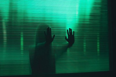 Foto einer Frau, die sich mit den Händen an einer grünen Glasfläche mit Lamellen abstüzt. Durch die Struktur der Glasfläche sieht man nur die Shilouette der Frau.
