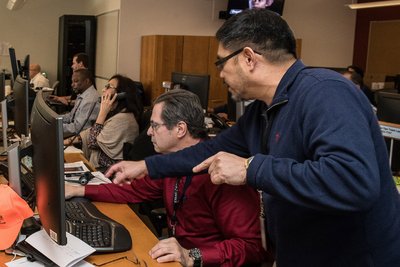 Foto von mehreren Mitarbeiter, die an ihren Computerarbeitsplätzen sitzen. Im Vordergrund steht ein Mann in angespannter Haltung, der auf etwas auf dem Monitor des Mannes neben ihm zeigt. 