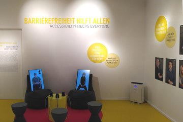 Photo of the Dialogue Lab station "accessibility helps all", where the visitor can sit on stools in fornt of two screens, which are placed on chairs, and watch interviews with persons with different disabilites. 