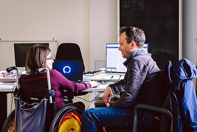 Foto von einer Frau im Rollstuhl und einem Mann, die gemeinsam an einem Computer arbeiten.