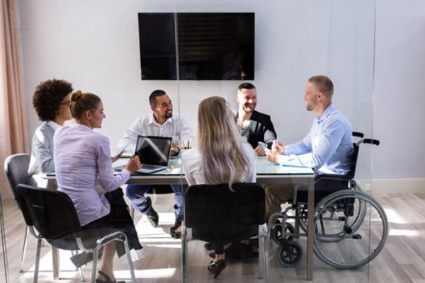 Foto einer Gruppe von Mitarbeitern, die zu einer Besprechung zusammen an einem Tisch sitzen. Einer von ihnen in einem Rollstuhl. 