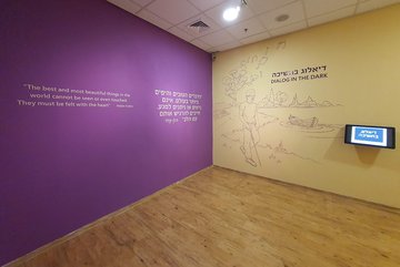 Photo of the lobby of Dialogue in the Dark Israel, a wall with a quote of Helen Keller: "The best and most beautiful things in the world, cannot be seen or even touched. They must be felt with the heart." in English and Hebrew.
